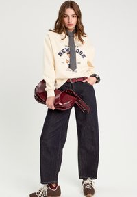 Sweatshirt beige avec un design imprimé, pantalon en denim faiblement taille haute de couleur foncée, sac bordeaux et baskets marron avec des lacets blancs. Accessoire cravate gris.