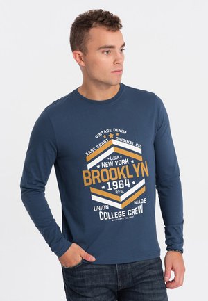 Camiseta de manga larga en color azul marino con estampado gráfico que presenta texto en naranja y blanco, cuello redondo y textura suave; combinada con jeans azules.