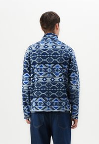 Blå og hvid mønstret fleece-pullover med høj krave, der har geometriske designs og en afslappet pasform, parret med mørke denimbukser.