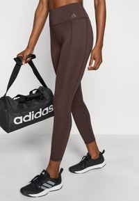 Leggings marron avec une taille haute, présentant un logo subtil et des détails de coutures, accompagnés d'un sac de sport noir et de chaussures de sport noires.