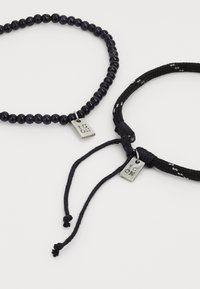 To sorte armbånd med små sølv "JEG ELSKER DIG" mærker, et perlearmbånd og et flettet armånd med justerbare snore, på en hvid baggrund.