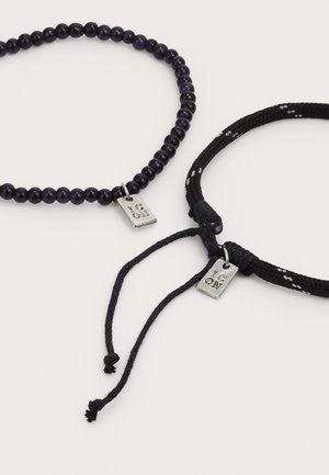 To svarte armbånd med små sølv "JEG ELSKER DEG" merker, ett perlearmbånd og ett flettet med justerbare snorer, på hvit bakgrunn.