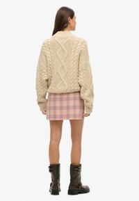 Beżowy sweter w splot kablowy z diamentowym wzorem, zestawiony z różową mini spódnicą w kratikę. Czarne botki za kostkę z paskami i klamrami.