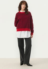 Un pull rayé rouge et marine avec un col rond, porté par-dessus une chemise blanche à col, associé à un pantalon large noir et des chaussures pointues noires.