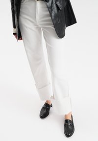 Witte, rechte jeans met een rafelige zoom, gecombineerd met zwarte instapschoenen met metallic accenten, tegen een effen witte achtergrond.