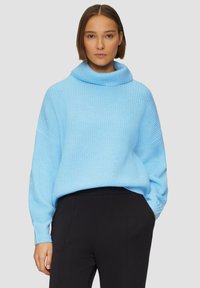 Lichtblauwe gebreide trui met een cowl neck, wijde mouwen en een relaxed fit. Gepaard met zwarte broeken, tegen een neutrale achtergrond.