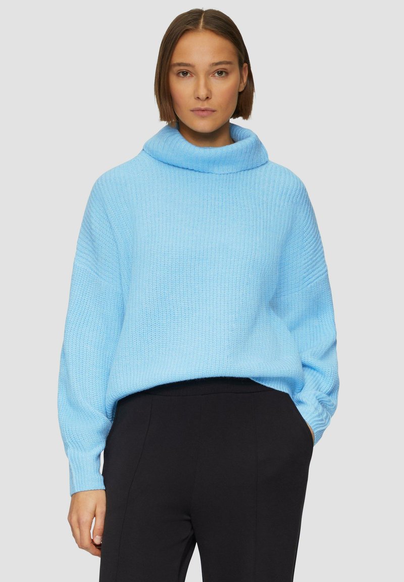 Lichtblauwe gebreide trui met een cowl neck, wijde mouwen en een relaxed fit. Gepaard met zwarte broeken, tegen een neutrale achtergrond.