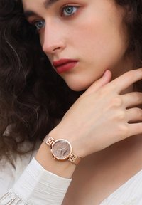 Orologio da polso in oro rosa con quadrante rotondo texturizzato, cinturino in rete metallica e accenti minimalisti in argento e oro rosa, indossato su un polso.