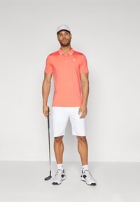 Polo corallo con rifiniture bianche, abbinato a pantaloni corti bianchi. Il modello tiene un bastone da golf, indossando un cappellino bianco e scarpe da golf grigie con accenti verdi.
