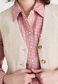 Gilet in velluto a coste beige con un design strutturato, caratterizzato da due tasche frontali e tre bottoni rotondi, indossato sopra una camicia a quadri rossi e bianchi.