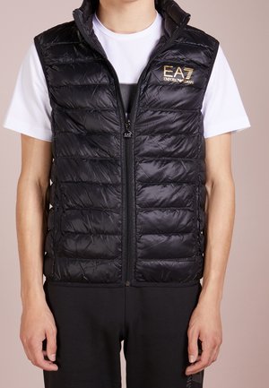 Czarna pikowana kamizelka zapinana na zamek z logo EA7 Emporio Armani, noszona na białej koszulce i czarnych spodniach na stojącej osobie.