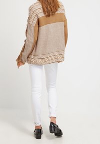 Sweter dzianinowy w beżowym kolorze z wzorzystym designem, z frędzlami. Połączony z białymi dżinsami i czarnymi butami, widok z tyłu.
