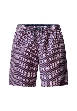 Hombre sin camisa de pie contra un fondo liso, vestido con pantalones cortos de baño estampados y zapatos beige sin cordones, mirando hacia adelante con una expresión neutral.