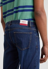 Mörkblå jeans med en figurnära design, med fem fickor och en vit etikett med röda accenter. En grönrandig skjorta är delvis synlig ovanför.