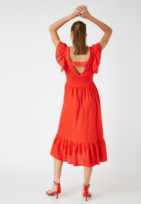 Vestido de lino rojo con mangas abullonadas, un escote cuadrado en la espalda y un dobladillo de volantes en capas. Se lleva con sandalias de tacón rojas a juego.