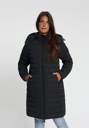 Vrouw met lang bruin haar die een lange zwarte gewatteerde winterjas met capuchon draagt, staand met handen in de zakken tegen een effen achtergrond.