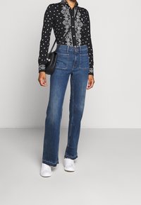 En person i en svart mönstrad långärmad skjorta, blå jeans med framfickor, vita sneakers och en svart axelväska.