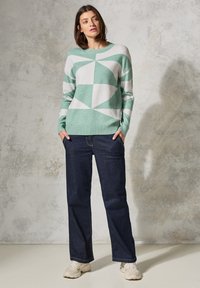 Grüner und weißer Pullover mit geometrischem Muster und Rundhalsausschnitt, kombiniert mit dunkelblauen Jeans und hellen Sportschuhen.