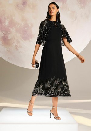 Schwarzes Midi-Kleid mit einer bestickten Kapuze, ausgestattet mit metallischen Akzenten und floralen Mustern am Saum. Getragen mit silbernen Riemchen-Absatzschuhen.