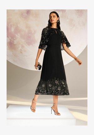 Schwarzes Midi-Kleid mit einer bestickten Kapuze, ausgestattet mit metallischen Akzenten und floralen Mustern am Saum. Getragen mit silbernen Riemchen-Absatzschuhen.