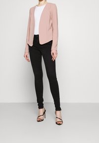 Blazer rose blush à coupe ajustée, manches longues, col pointu et devant ouvert. Porté sur un top blanc, associé à un jean skinny noir.