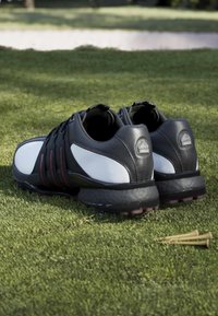 Pair of black and white golf shoes resting on green grass with three wooden golf tees nearby.