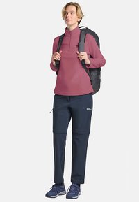 Fleece-pullover in lichtroze met een kwart rits, gecombineerd met marineblauwe stretchbroek met cargozakken. Zwarte rugzak en blauwe hardloopschoenen.