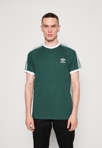 Žalias medvilninis marškinėliai su balta apykakle ir rankovių akcentais, su "Adidas" logotipu kairėje krūtinės pusėje ir dryžuota pečių dizainu.