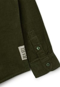 Camisa de pana verde oliva con puño abotonado, que presenta una etiqueta tejida con texto y una textura estampada en la tela.