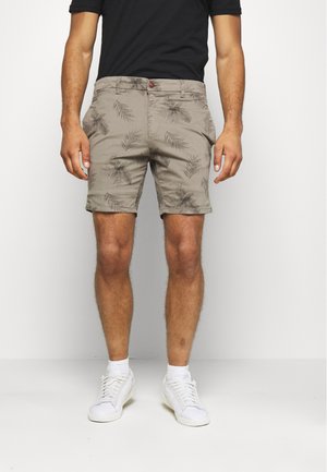 Homme portant un short beige à imprimés de feuilles foncées, associé à un t-shirt noir et des baskets blanches, debout devant un fond uni.