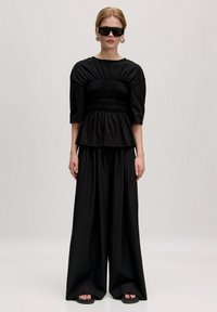 Zwarte peplumtop met geplooide details, gecombineerd met wijd uitlopende zwarte broeken en platte sandalen. Accessoires omvatten oversized zwarte zonnebrillen.