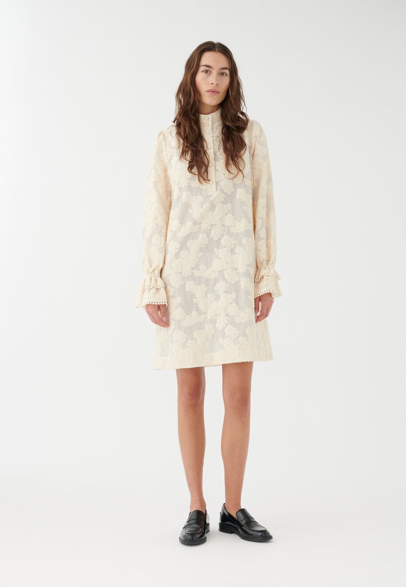 Vestido de encaje en crema con patrón floral y cuello alto. Mangas largas con puños con volantes. Combinado con mocasines negros.