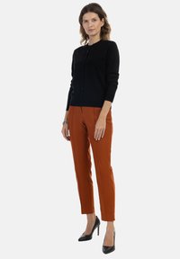 Czarny kardigan z guzikami, zestawiony z wąskimi spodniami w kolorze rdzy. Modelka nosi czarne szpilki. Prosty, elegancki design.