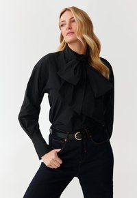 Camicione nero a maniche lunghe con un grande colletto a farfalla, texture liscia e vestibilità strutturata. Abbinato a jeans scuri e una cintura nera.