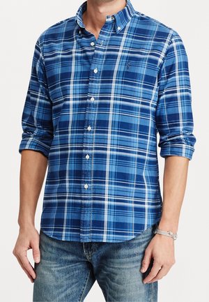 Mężczyzna w niebiesko-granatowej koszuli w kratę z podwiniętymi rękawami i wyblakłych niebieskich dżinsach, stojący z rękami luźno opuszczonymi wzdłuż ciała.