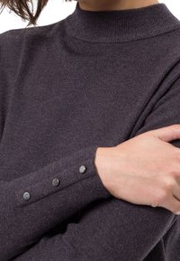 Haut à manches longues violet foncé avec un col haut et trois boutons argentés sur chaque poignet. Texture tricot doux, silhouette ajustée.