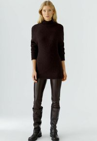 Donkerbruine gebreide trui met een hoge hals, voorzien van lange mouwen en een relaxte pasvorm, gecombineerd met glanzende bruine leren leggings en hoge laarzen.