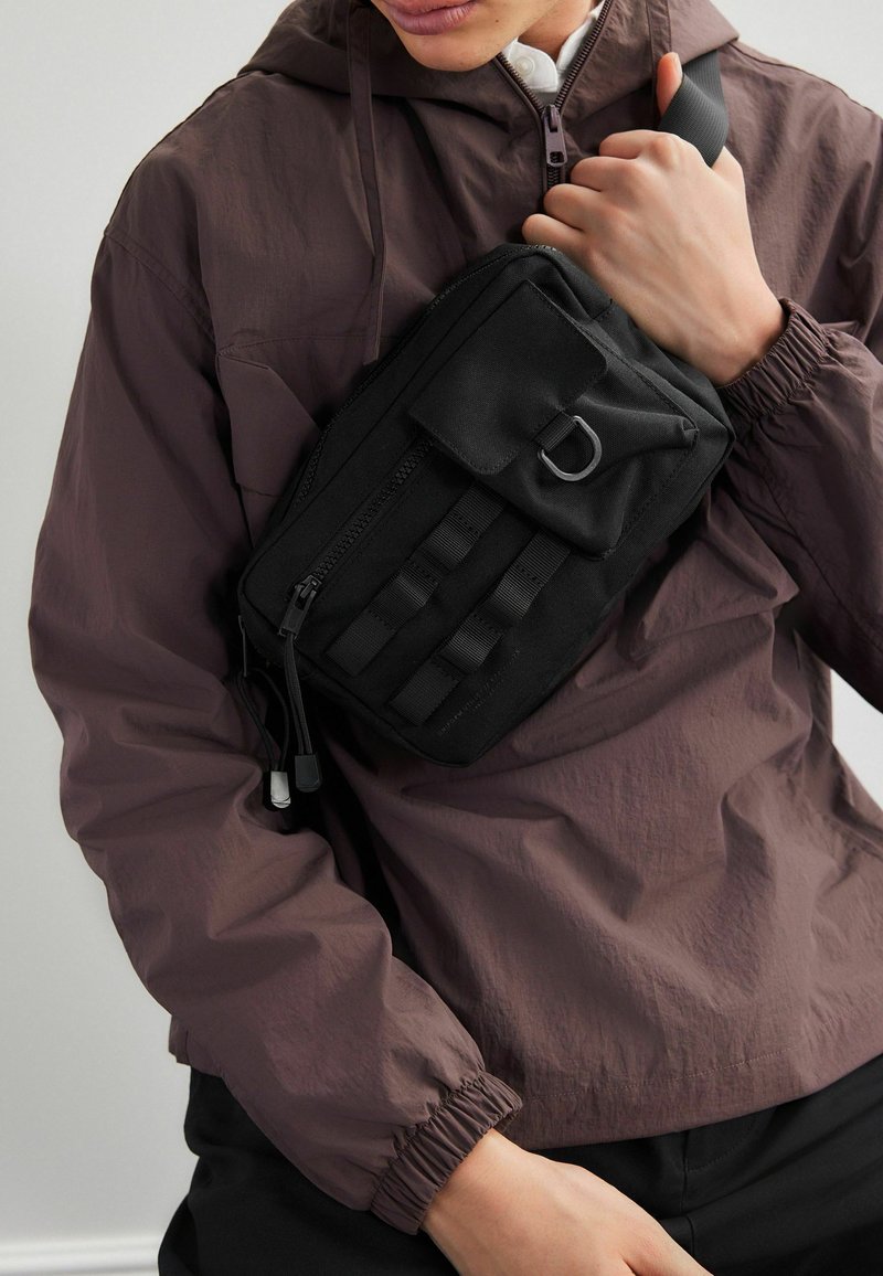 Black shoulder bag made of durable fabric, featuring a flap, zippered compartments, and vertical webbing straps for attachment.