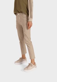 Pantaloni beige alla caviglia con tasche frontali e una sottile trama, abbinati a sneakers beige chiaro con suola bianca e dettagli dorati.