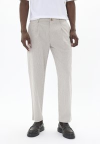Pantalones rayados de color beige claro hechos de tela texturizada; con plisados frontales y un cierre de un solo botón. Combinados con zapatos negros tipo slip-on.