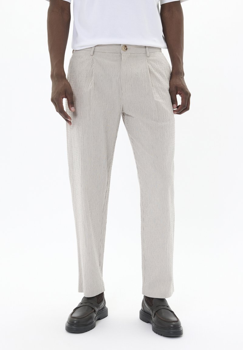 Pantalones rayados de color beige claro hechos de tela texturizada; con plisados frontales y un cierre de un solo botón. Combinados con zapatos negros tipo slip-on.