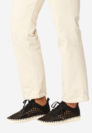 Voeten in zwarte mesh veterschoenen met witte geribbelde zolen en crèmekleurige broek tegen een effen achtergrond.