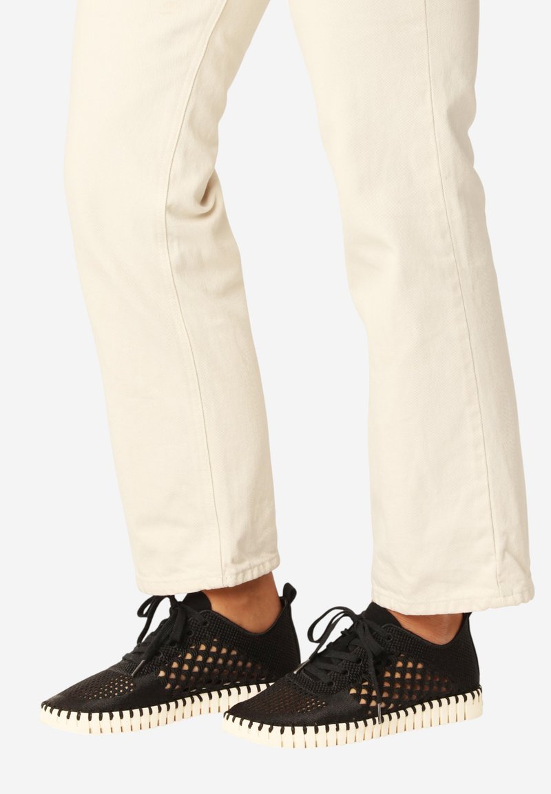 Voeten in zwarte mesh veterschoenen met witte geribbelde zolen en crèmekleurige broek tegen een effen achtergrond.