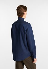 Camicia in denim blu scuro con colletto a bottoni, maniche lunghe e orlo curvo. Tessuto liscio con cuciture tonali e dettagli con bottoni.