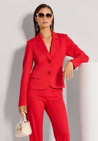 Rode op maat gemaakte pak met een aansluitende blazer en pantalon. De blazer heeft geknipte revers en twee knopen aan de voorkant. Accessoires omvatten grote zonnebrillen en een kleine handtas.