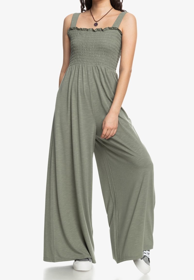 Combinaison en tissu vert avec jambes larges, corsage froncé et col à volants. Comprend des bretelles et une texture lisse.