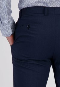 Man met donkerblauwe op maat gemaakte broek en een lichtgepatroneerd overhemd, met één hand in de zijzak van de broek.