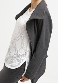 Schwarze, strukturierte Jacke mit weitem Kragen, darüber ein helles, gemustertes weißes Top. Das Modell posiert seitlich und präsentiert das Ensemble.