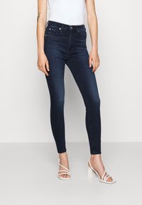 Dunkelblaue Skinny-Jeans aus Denim mit mittelhohem Bund, Vordertaschen und nahtlosem Design. Kombiniert mit weißen Absatzsandalen.