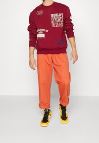 Felpa color marrone scuro con varie stampe, abbinata a pantaloni arancioni ampi e sneaker nero-gialle con dettagli bianchi.